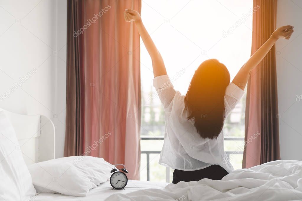 Happy asian woman stretching on bed after waking up at morning in bedroom with sunlight shine through curtains