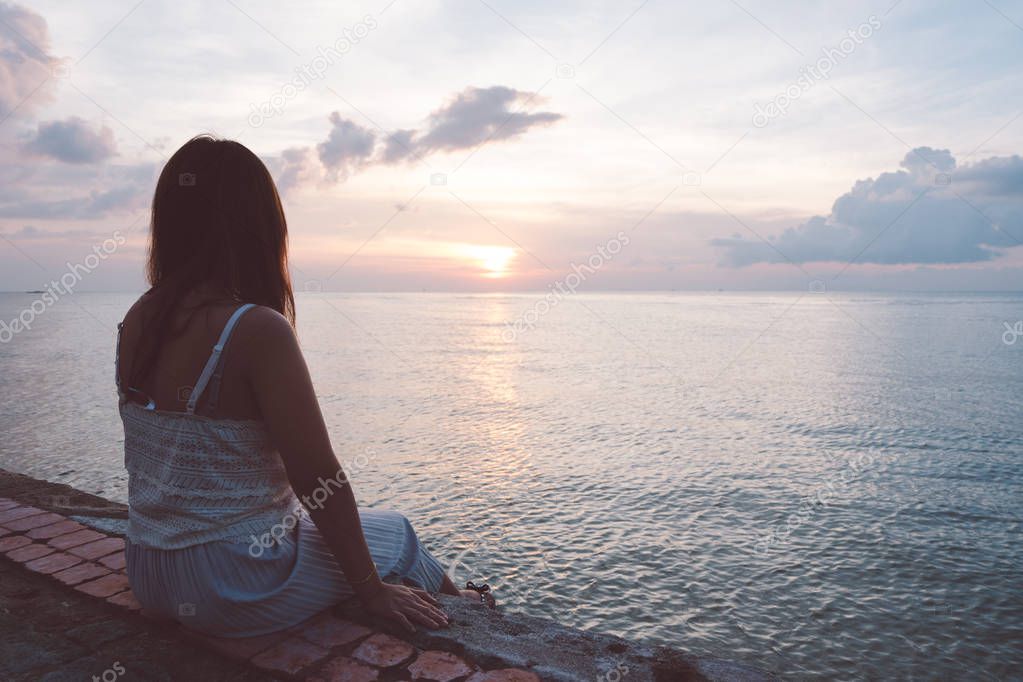 Silhouette of young woman sitting alone on back side outdoor at tropical island beach missing boyfriend and family in summer sunset. Sad and lonely concept in dark and vintage tone.