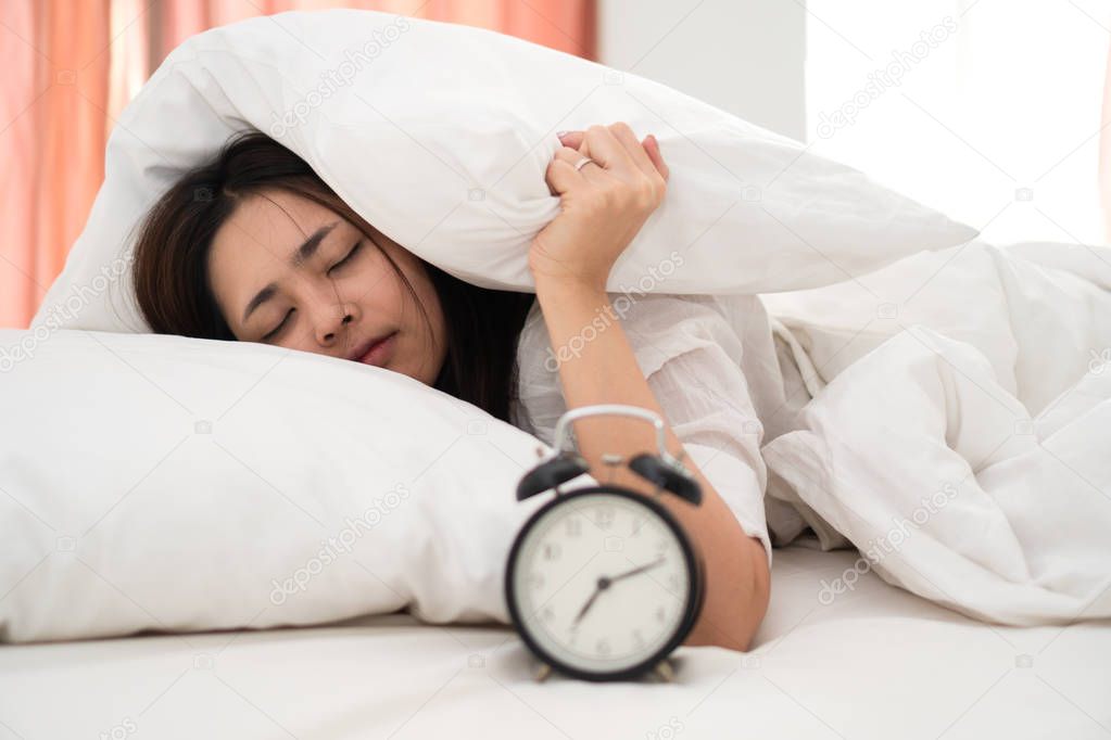 Young asian woman lazy waking up in early morning for routine daily job. Sleepy girl lying on bed closing her eyes and try to hiding under pillow for avoid alarm clock snooze.