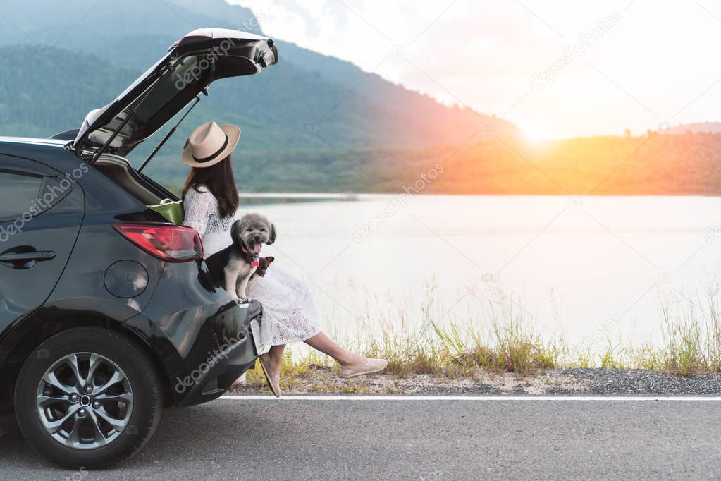 Happy young woman traveler sitting in hatchback car with dogs at lake, mountain view and sunset. Cheerful female in white dress travels with pets in summer vacation in road trips. Outdoor relaxation.