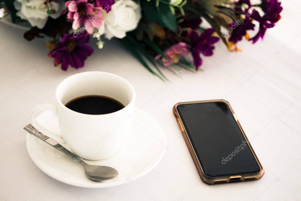 White cup of black coffee with blank smartphone screen and flower placed on white table background at coffee shop in fresh morning. 