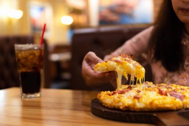 Slice of hot pizza with stringy cheese in hand. Young woman enjoy eating Hawaiian pizza topped with tomato sauce, cheese, pineapple and bacon on wooden board with soft drink in restaurant for dinner clipart