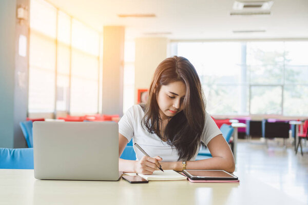 Attractive young asian female student studying ebook online course and writing notes on notebook in library while preparing for exam with laptop and tablet pc, Learning and education concept