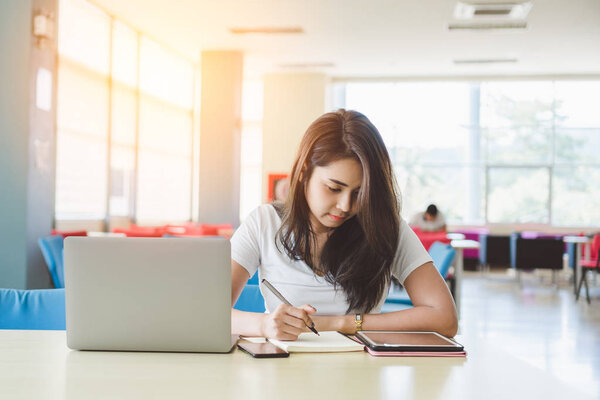 asian female student writing on notebook while sitting at table with  laptop and tablet pc  in library,  education and research concept