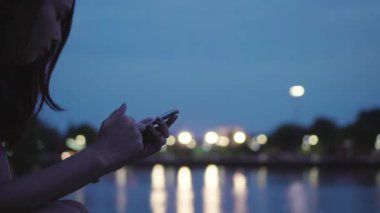 Su Nehri yakınında oturan sokak ışıkları ile gece sırasında cep telefonuyla çekici Asyalı kadın yansıyan.