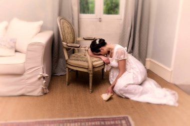 Depressend youg woman in authentic regency dress holding a letter and leaning against an antique chair clipart
