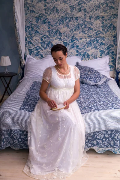 有吸引力的年轻女子在正宗的摄政礼服坐在古色古香的床上 — 图库照片