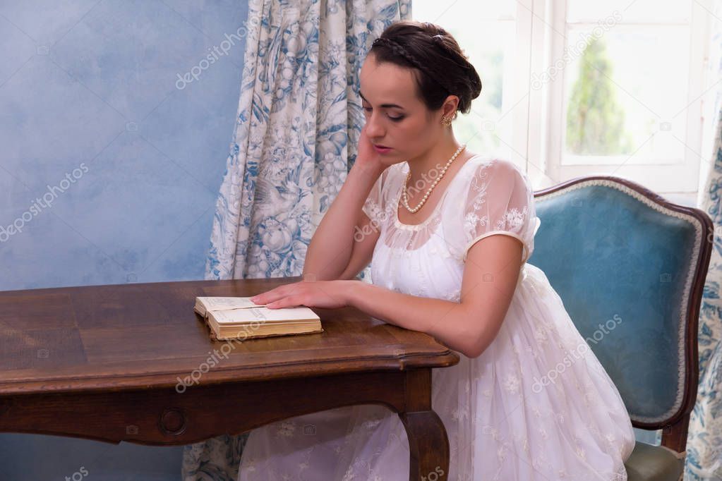 Pretty young woman in authentic regency dress reading a book in vintage room