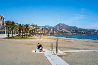 Malaga, İspanya, Nisan 2016: Yazın güneşli ve açık gökyüzü olan La Malagueta plajı manzaralı. Palmiye ağaçları, şehir mimarisi ve arka planda dağlar