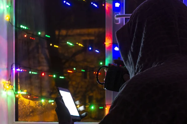 sad man in a hood with a smartphone in a blurred bokeh, on the background of the window decorated with garlands with an empty glass and a bottle of champagne, in anticipation of the new year and Christmas