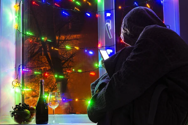 sad man in a hood with a smartphone in a blurred bokeh, on the background of the window decorated with garlands with an empty glass and a bottle of champagne, in anticipation of the new year and Christmas