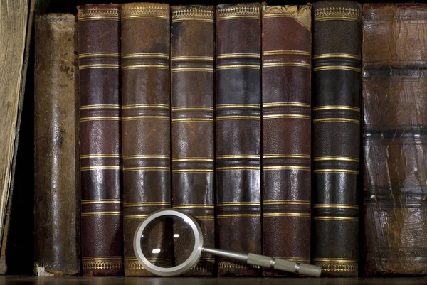 background: a row of worn leather antiquarian book spines with gold embossing and a magnifying glass