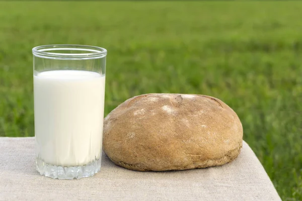牛乳とぼやけ草原の背景にプレーン リネン テーブル クロスとテーブルの隅に素朴なパンのガラス — ストック写真