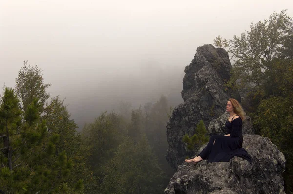 红头发的赤脚女孩穿着黑色连衣裙 坐在森林上方的岩石上 面对雾的临近墙 岩石是山 Kolpaki 的顶峰和欧洲和亚洲在中部乌拉尔的条件边界 — 图库照片