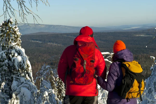 The man and woman hikers stand with their backs to the viewer at the top of the mountain in winter and look into the distance
