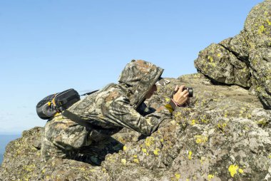 girl wildlife photographer in camouflage suit in the mountains hiding behind a stone photographing something clipart