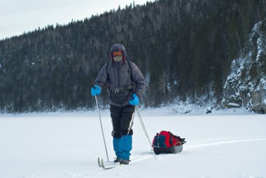 rocky, woody kıyı karşı bir donmuş nehir boyunca yürüyüş bir kargo kızak ile kayakçı