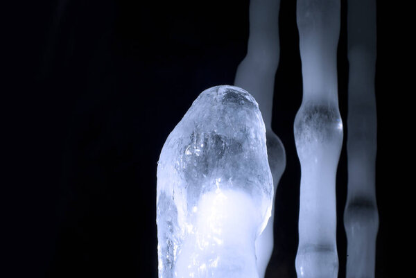 верхняя часть пещерного ледяного сталагмита крупным планом на черном с несколькими размытыми другими сталагмитами
 