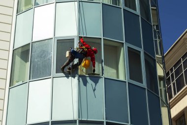 rope access technician in a helmet washes the window of a high-rise building from the outside, hanging on a rope clipart