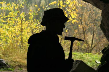 silhouette of a geologist with a geological hammer in a cave, on the background of an entrance with a brightly lit autumn forest clipart
