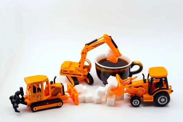 toy construction machines loads cube sugar into a real cup of coffee on a light background