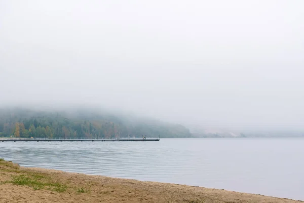 Shore of an autumn lake in fog and a pier in the distance — Stock Photo, Image