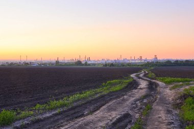 Akşam kırsalında tarlalar, toprak yol ve ufukta büyük bir petrol rafinerisi var.