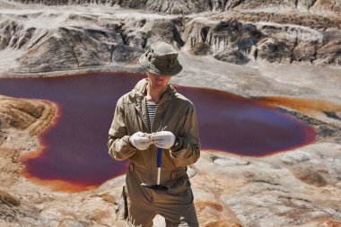 Jeolog, kırmızı tuz gölü olan bir kanyonun arka planında bir mineral örneğini inceliyor.
