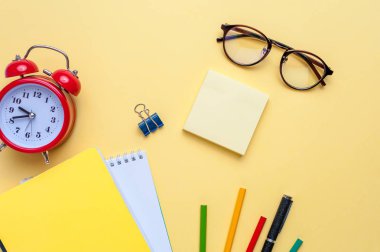 Çalar saat, boş defter, sarı arka planda kalem.