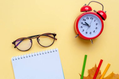 Çalar saat, boş defter, sarı arka planda kalem.