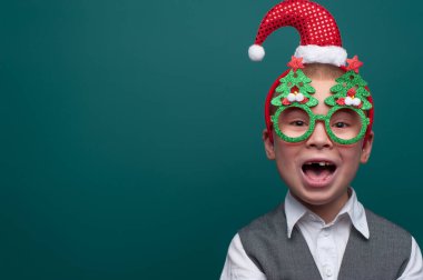 Noel Baba şapkalı neşeli çocuk portresi ve Noel ağaçlı komik gözlükler.