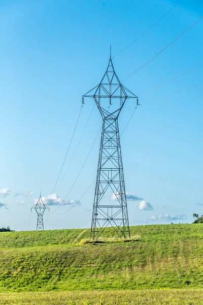 Güneşli bir günde düz ve yeşil bir tarım arazisinde yüksek voltaj hatları ve elektrik direkleri.