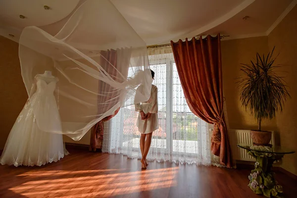 The bride in a dressing gown near the window. Waves veil.