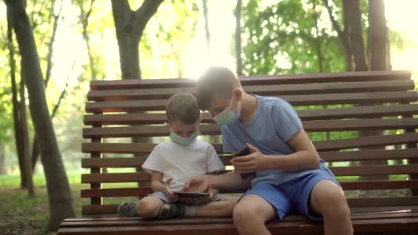 Niños con máscaras médicas juegan con smartphones en el Parque — Vídeos de Stock
