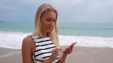 Sahilde duran ve deniz fırtınası vasıl geçmiş üzerinden smartphone kullanarak siyah ve beyaz elbiseli uzun sarı saçlı güzel ince kadın... Kız sahilde ekran ve gülümseme dokunmadan.