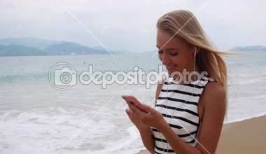 Sahilde duran ve deniz fırtınası vasıl geçmiş üzerinden smartphone kullanarak siyah ve beyaz elbiseli uzun sarı saçlı güzel ince kadın... Kız sahilde ekran ve gülümseme dokunmadan.