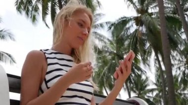 Siyah ve beyaz elbiseli uzun sarı saçlı güzel ince kadın onun telefon görünüyor, kız bir telefon bir ayna yerine kullanır ve saçlarını ayarlar. Curvy saç.