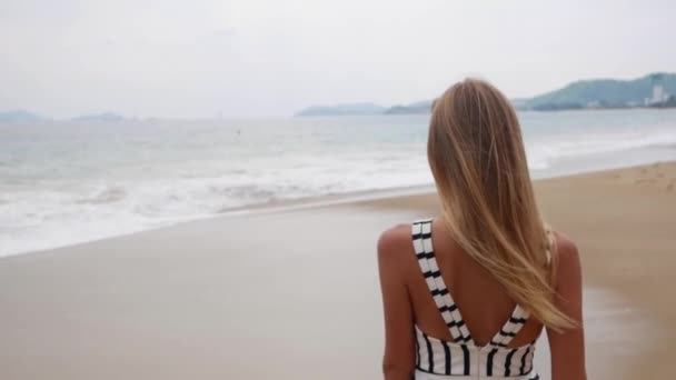Leende kvinna i svart och vitt klänningar går ensam på en tropisk strand och tittar på kameran över bakgrunden på storm på havet. — Stockvideo
