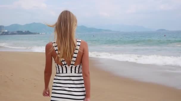 Jovem mulher sorridente em vestidos pretos e brancos andando sozinha em uma praia tropical e olhando para a câmera sobre o fundo na tempestade no mar . — Vídeo de Stock