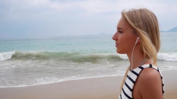 Jeune belle femme mince avec de longs cheveux blonds en robe noire et blanche avec écouteurs et écoute de la musique marchant sur fond de tempête sur la mer . — Video