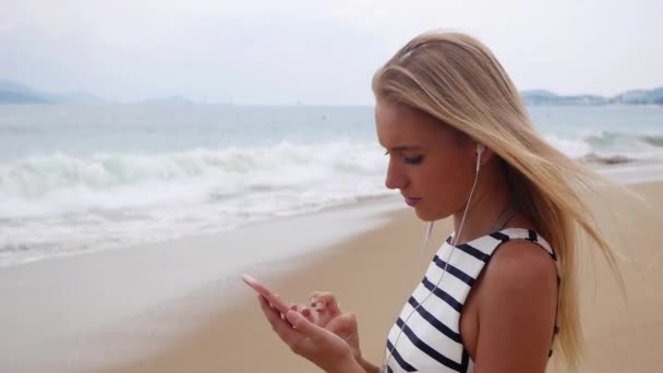 Schöne schlanke Frau mit langen blonden Haaren in schwarz-weißem Kleid an der Küste stehend und mit Smartphone über dem Hintergrund bei Sturm auf dem Meer.. Mädchen am Strand berühren Bildschirm und lächeln. — Stockvideo