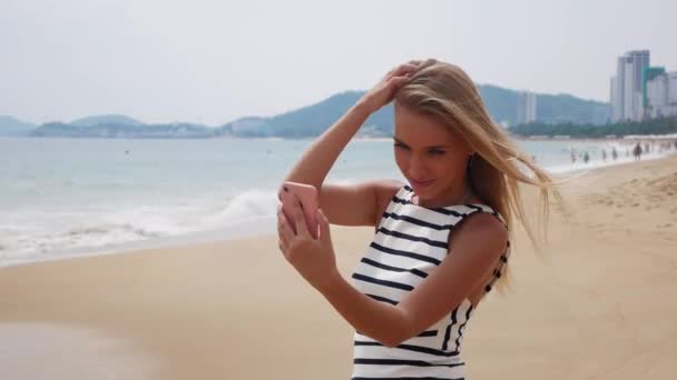 Mooie slanke vrouw met lang blond haar in zwart / wit jurk selfie maken op mobiele telefoon in de buurt van de zee op de achtergrond van de stad — Stockvideo