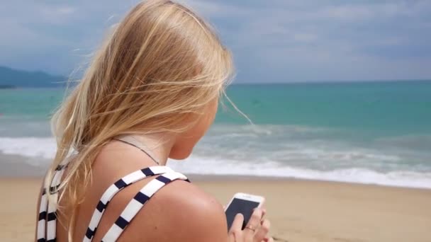 Schöne Frau schlank mit langen blonden Haaren in schwarz-weißem Kleid sitzt am Strand und benutzt ein Smartphone. Mädchen am Strand berührt den Bildschirm. — Stockvideo