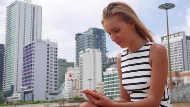 Hermosa mujer delgada con el pelo largo y rubio en vestido blanco y negro está sentado en la playa y el uso de un teléfono inteligente en el fondo de la ciudad. Chica en la playa tocando la pantalla . — Vídeo de stock