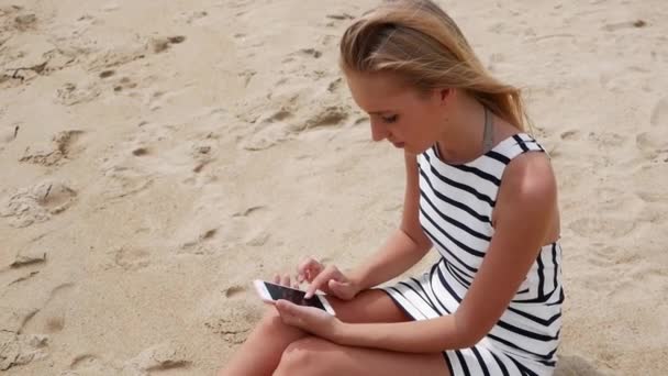 Beautiful woman slim with long blond hair in black and white dress is sitting on the beach and using a smartphone. Girl on the beach touching the screen. — Stock Video