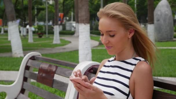 Piękne, szczupła kobieta z długie blond włosy w czarno-białej sukni siedzi na ławce i przy użyciu smartfona na tle parku. Dziewczyna na placu, dotykając ekranu i uśmiech. — Wideo stockowe