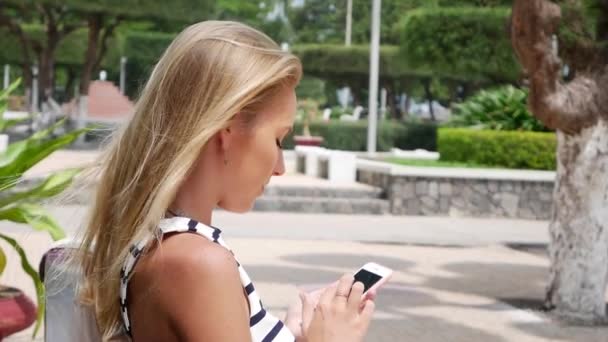 Schöne schlanke Frau mit langen blonden Haaren in schwarz-weißem Kleid sitzt auf der Bank und mit dem Smartphone über dem Hintergrund des Parks. Mädchen auf dem quadratischen Touchscreen. — Stockvideo