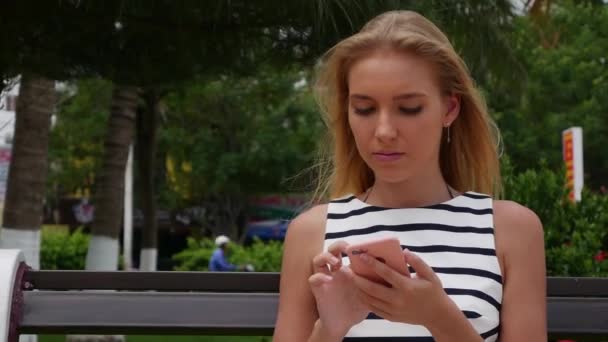 Schöne schlanke Frau mit langen blonden Haaren in schwarz-weißem Kleid sitzt auf der Bank und mit dem Smartphone über dem Hintergrund des Parks. Mädchen auf dem quadratischen Touchscreen. — Stockvideo