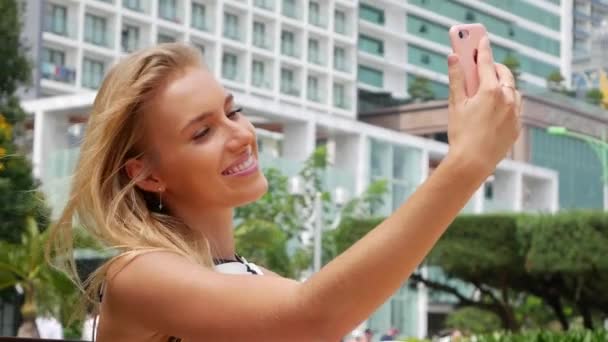 Hermosa mujer delgada con pelo largo y rubio en vestido blanco y negro haciendo selfie en el teléfono móvil sobre el fondo del parque . — Vídeo de stock