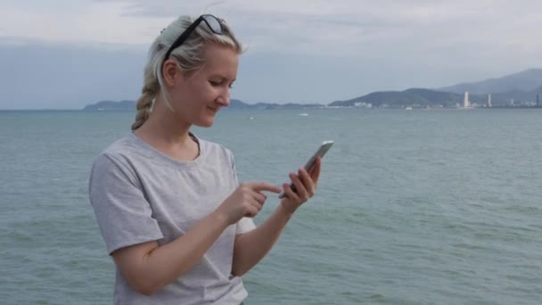 Schlanke Frau mit blondem Zopf im grauen T-Shirt, die mit dem Smartphone über dem Meer und den Inseln steht. Touchscreen für Mädchen — Stockvideo